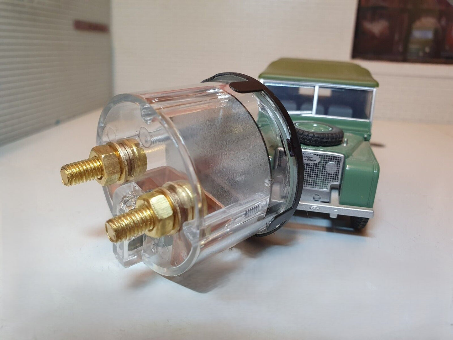 Rear view of an Ammeter showing two brass coloured screws and nuts and washers. To the right behind is a green Land Rover Series 1 used for illustration purposes.
