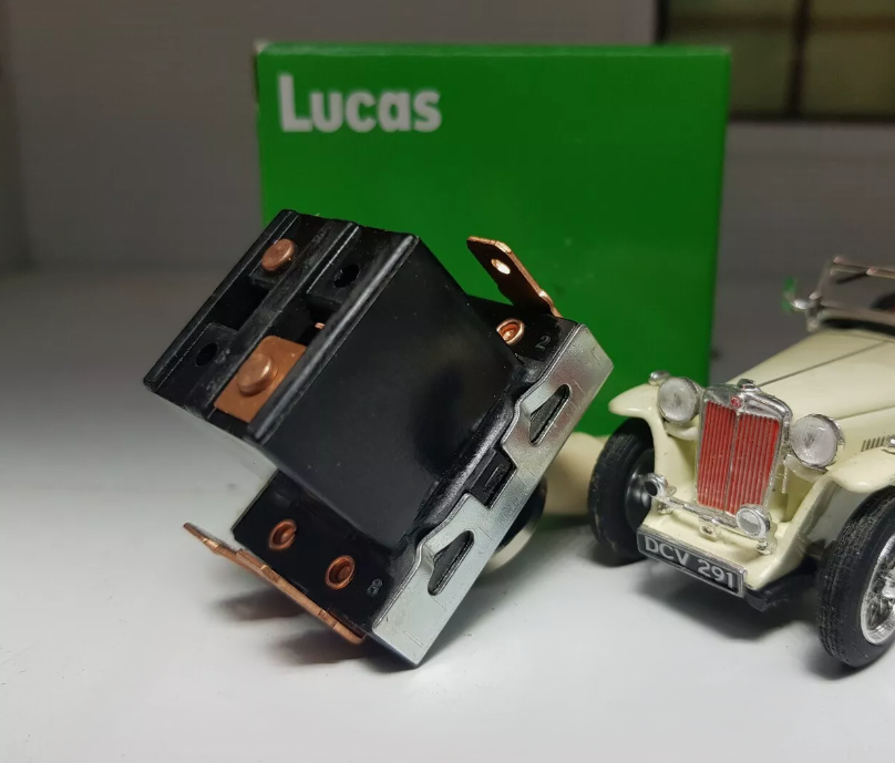Underside view of a toggle switch showing the terminals, in the background is a Green Lucas Box and to the right, a model of a classic car for illustrative purposes.