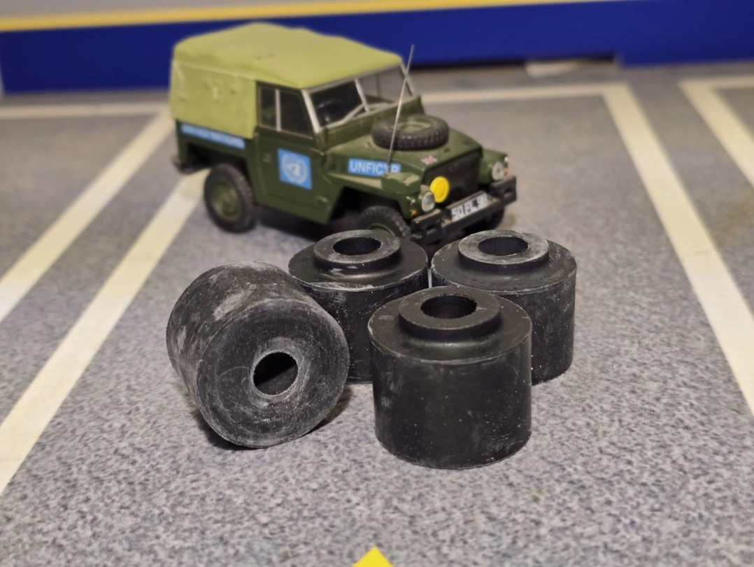 Four black cylindrical Tie Rod Bush displayed in the foreground on a gray asphalt surface, with a green vintage Land Rover model in the background.