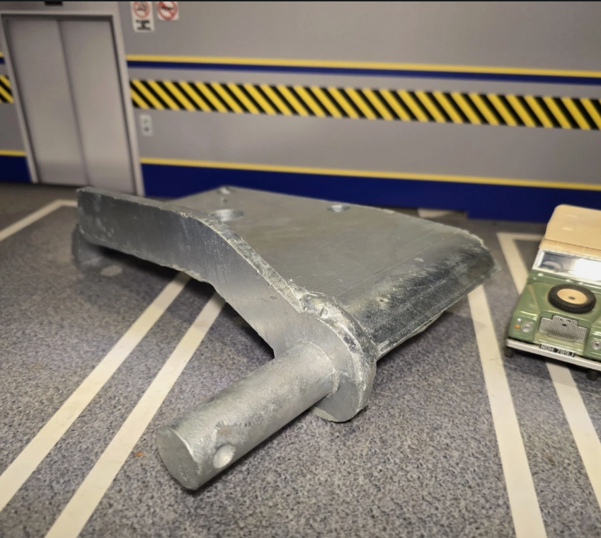 A large galvanized steel mounting bracket with a cylindrical bolt shaft, shown on a gray floor with white parking-style lines. A small green vintage model car is placed behind it for scale.