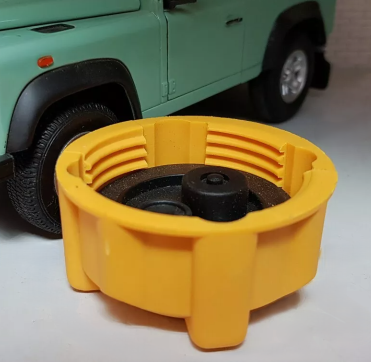 A yellow expansion tank cap, upside down, showing the threads and seal, on a grey surface with a heritage green defender model in the background (model for illustration purposes).