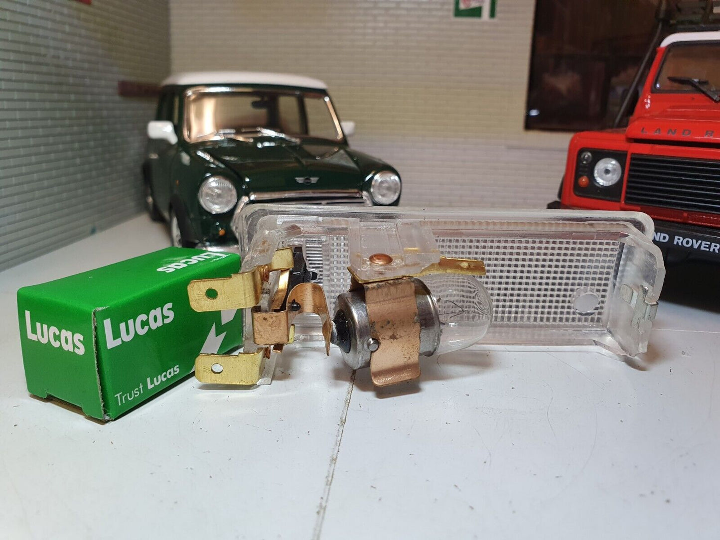 Rear view of an interior roof light, showing the bulb and inner workings. A Lucas box sits to the left. Behind To the left is a model of a classic green Mini, to the right is a model of a Red Land Rover Defender (models for illustration purposes only).