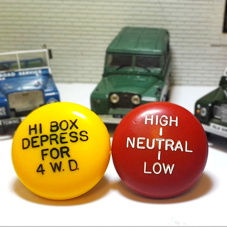 Two Shift Knobs, one yellow and one red, with text on a surface with model vehicles in the background.