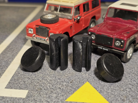 Two black rubber buffers and hinge bushes arranged on a toy road surface in front of two red Land Rover toy vehicles.
