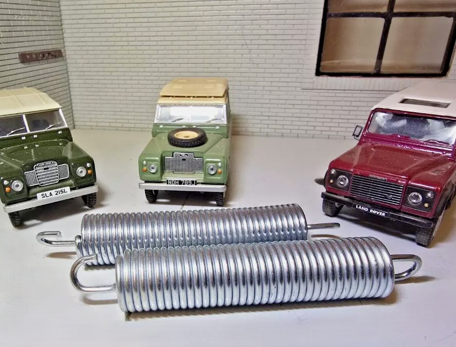 A Pair of pedal box return springs, on a grey surface, behind are model Land Rovers for illustration purposes.