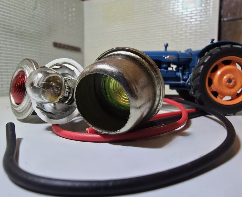 Red and Green Warning Lights with side by side with silver bezels and wiring tails. The green warning light has been disassembled to show the inside of the lens and the bulb and bulb holder, In the background is a Fordson Tractor for illustration purposes.