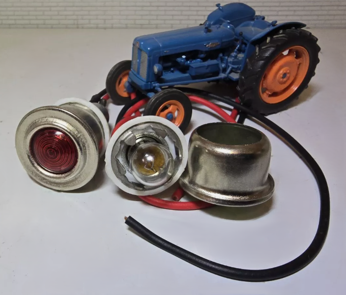 Red and Green Warning Lights with side by side with silver bezels and wiring tails. The green warning light has been disassembled to show the bulb and bulb holder, In the background is a Fordson Tractor for illustration purposes.