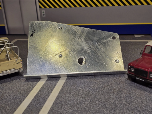 A brushed silver metal plate or bracket with pre-drilled holes, photographed on a gray tabletop with toy cars partially visible at the left and right edges.
