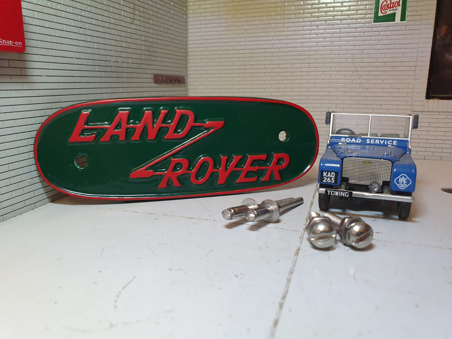 Red and green variant of a pressed aluminium Land Rover Series 1 grill badge, on the right is the model Series 1 (for illustration purposes only.) In front of the model are stainless rivets and domed headed slot machine screws for fitting.