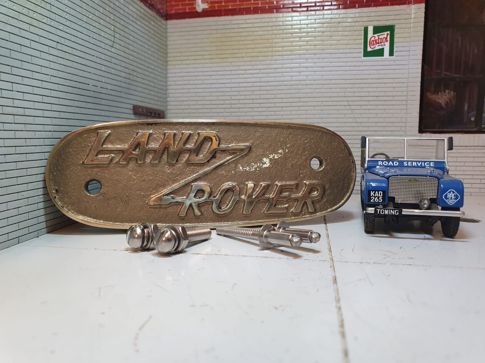 Brass variant of a pressed aluminium Land Rover Series 1 grill badge, on the right is the model Series 1 (for illustration purposes only.) In front of the badge are stainless rivets and domed-headed slot machine screws for fitting.
