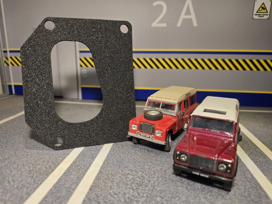 A black textured gasket with a large oval cutout stands next to two small toy cars in a miniature parking-lot style display. The background shows gray floor markings, a gray wall with blue and yellow hazard stripes, and the number 2A.