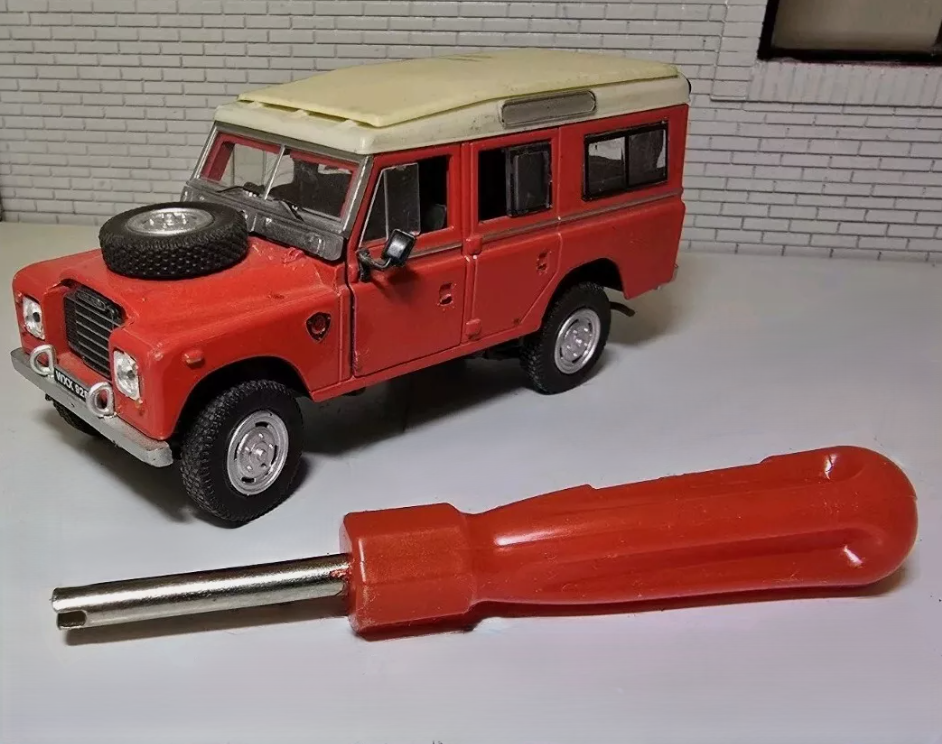 A red handled valve removal tool on a grey surface. To the back left of the image is a model of a red series 3 station wagon for illustrative purposes.