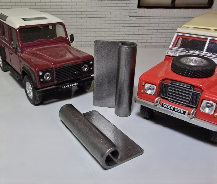 A Pair of Bulkhead Air Vent Flap Hinge Brackets, one in the foreground is laid down, the other is stood on end. To the left and right are Red Land Rover Defender and Series Models for Illustration Purposes.