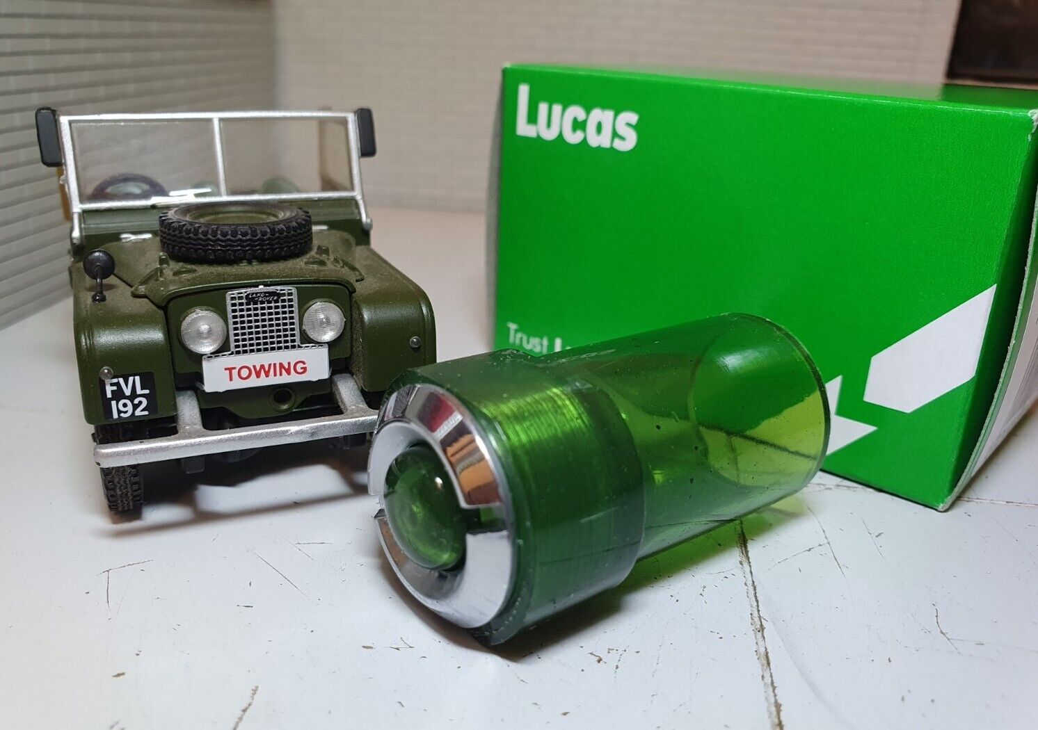 3/4 View of a Green Warning Light With Chrome Bezel with A Model Land Rover and Lucas Box in the Background